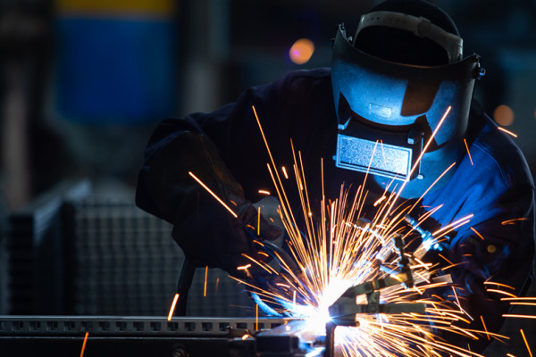 Person in a factory wearing safety gear welding, while sparks fly.